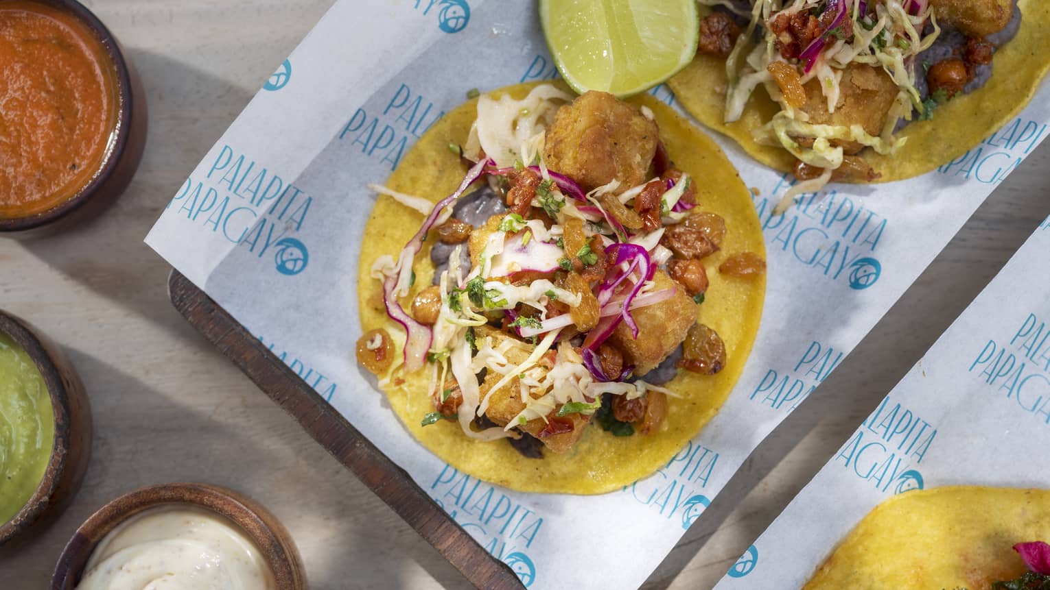 Falafel topped with lettuce, red cabbage and yogurt sauce served on top of a corn tortilla with three small bowls of sauces on the side