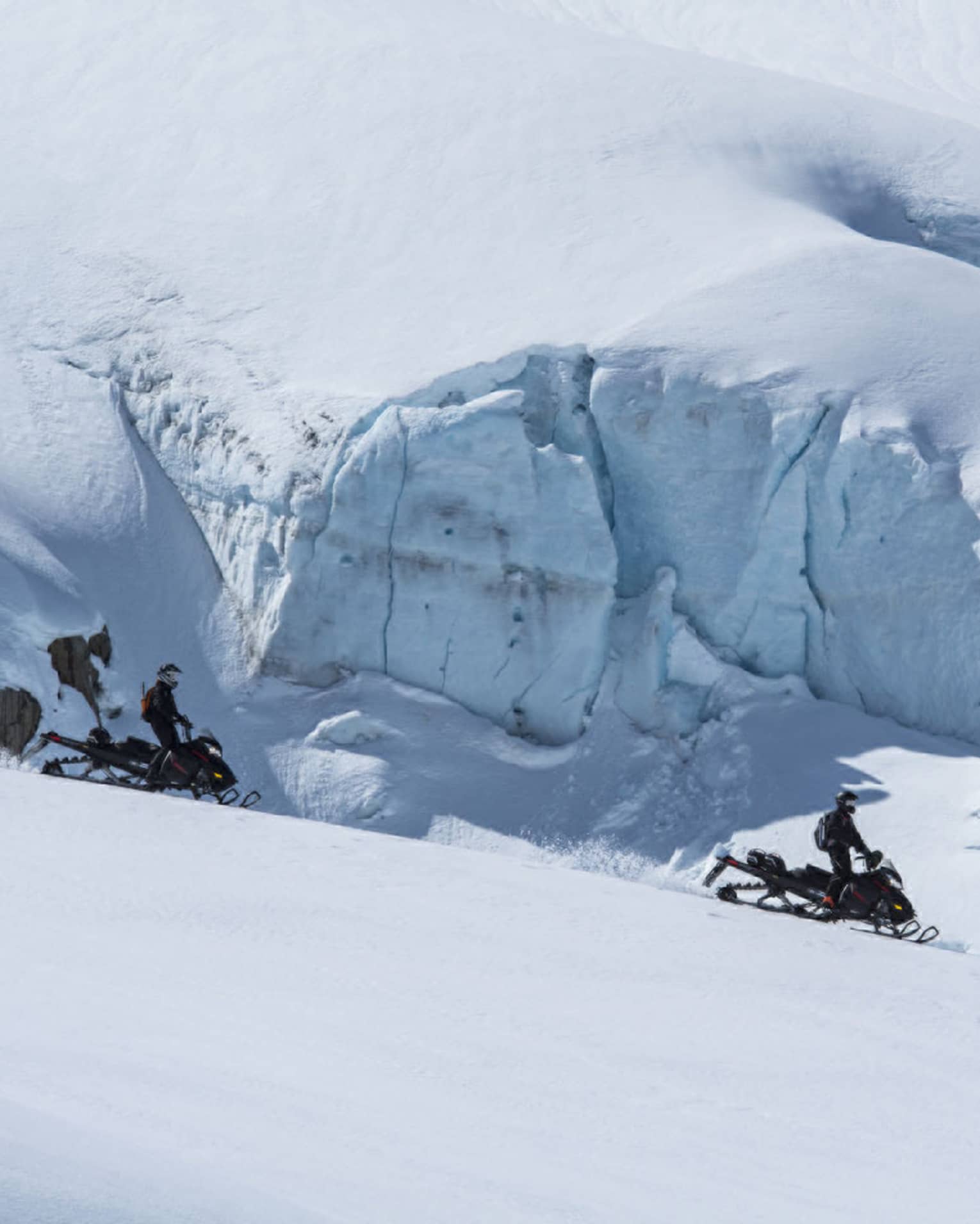 Side view of two distant black-clad snowmobilers coasting down a white slope alongside an enormous wall of ice and snow.