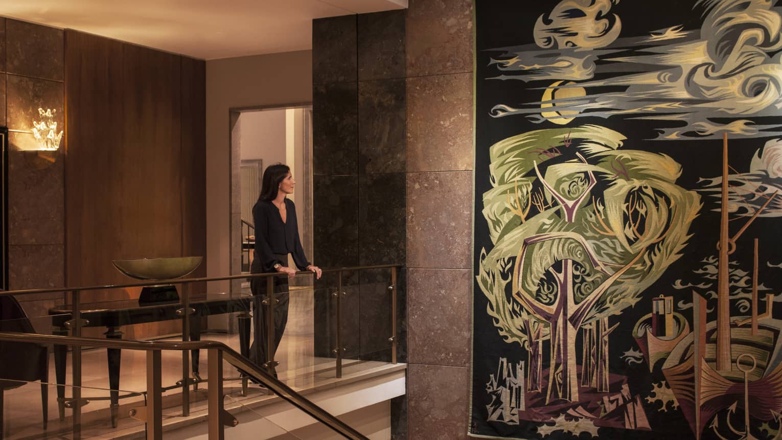 Woman dressed in black stands at glass railing in hotel lobby, looks at large colourful tapestry