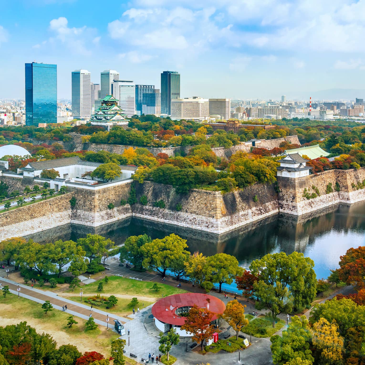 Osaka Castle rises amid lush autumn-coloured trees within a stone fortress surrounded by a moat and modern high-rises.