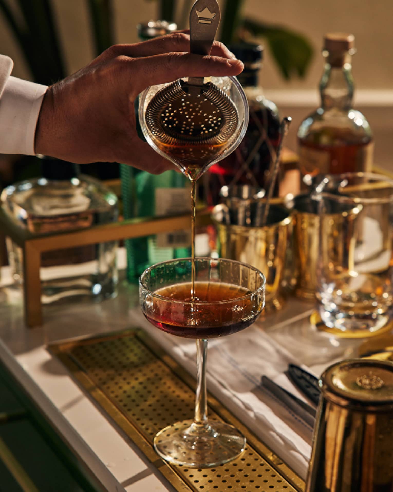 A bartender pouring a drink into a cocktail glass with bottles of liquor around.