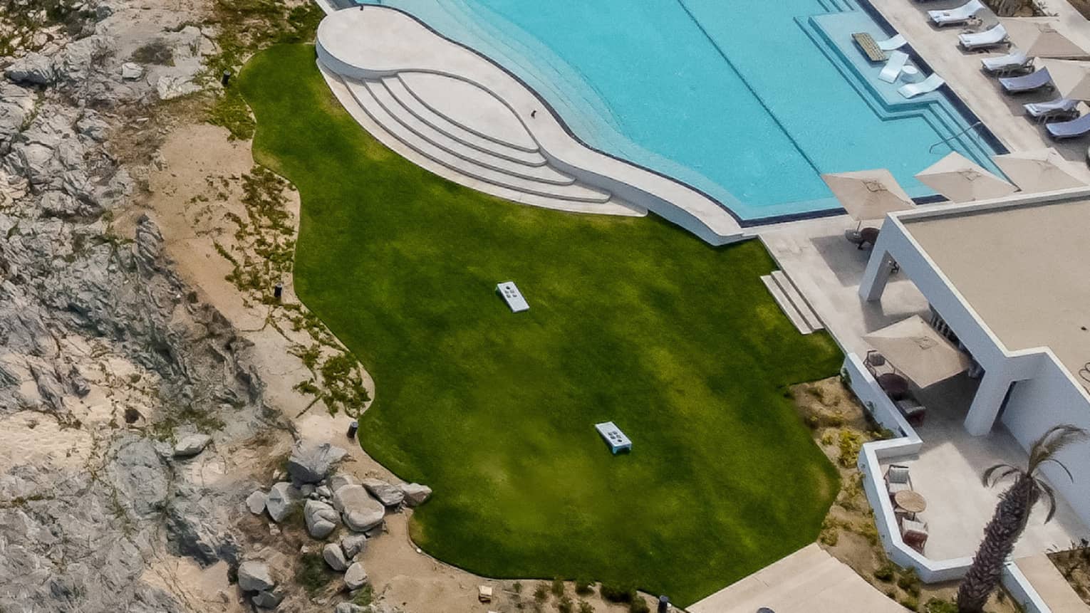 A bird's-eye view of a resort's grounds and beach next to the ocean shore. There is a large pool and event lawn space positioned between the resort and the beach.