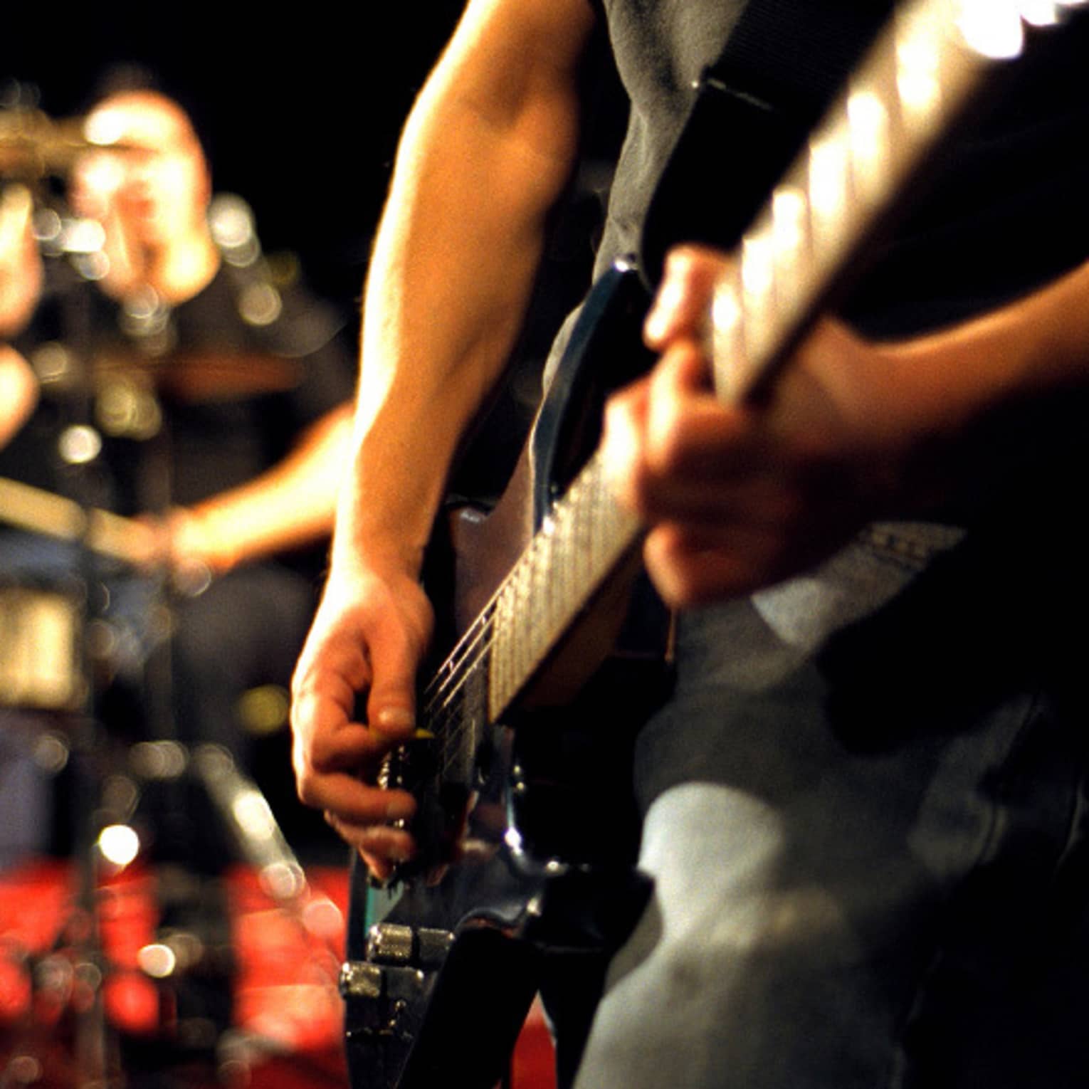 Close-up of musician's arms and hands playing an electric guitar with drumset in the background.