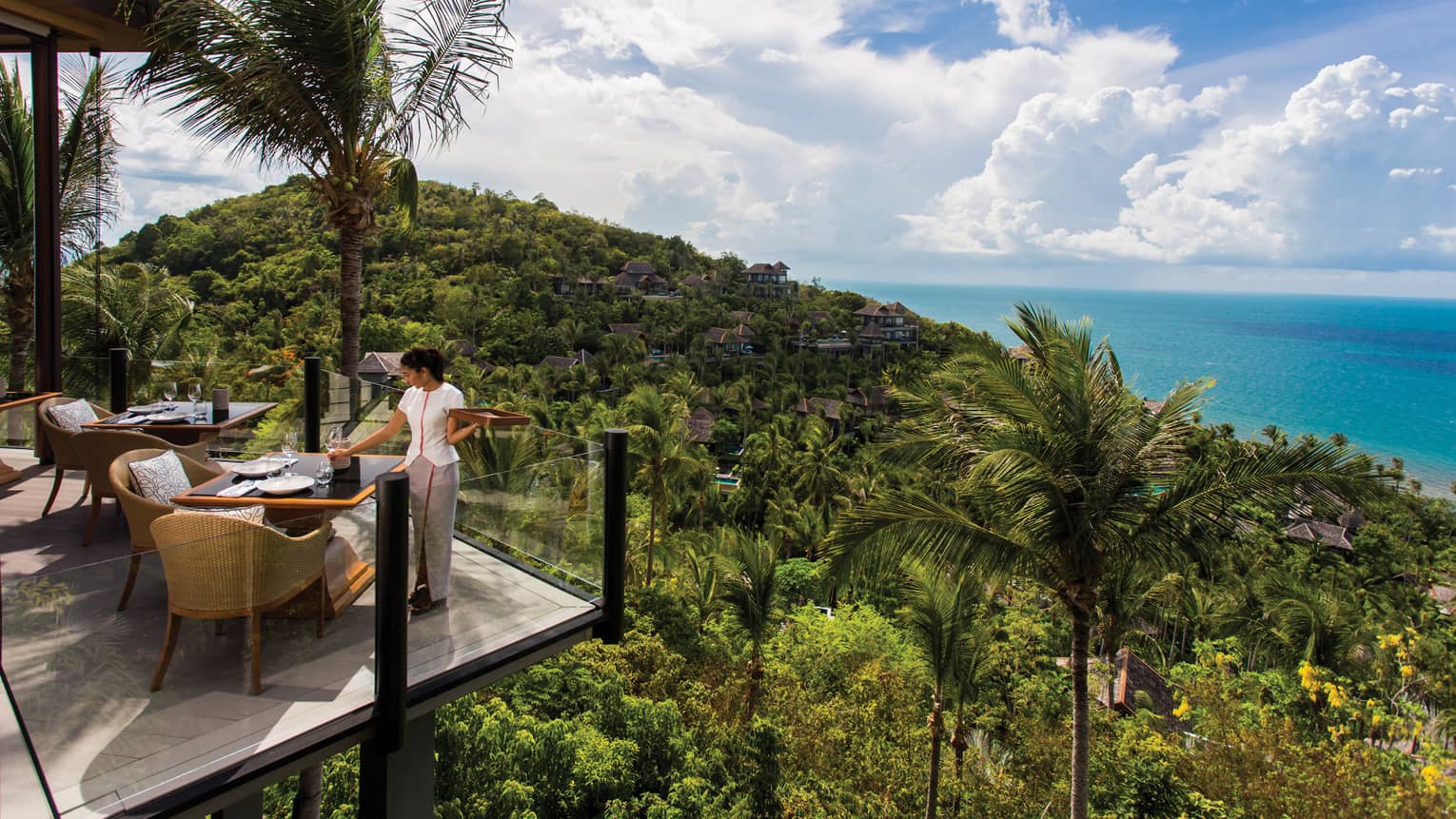 Server sets corner patio table behind glass balcony, high on tropical mountain