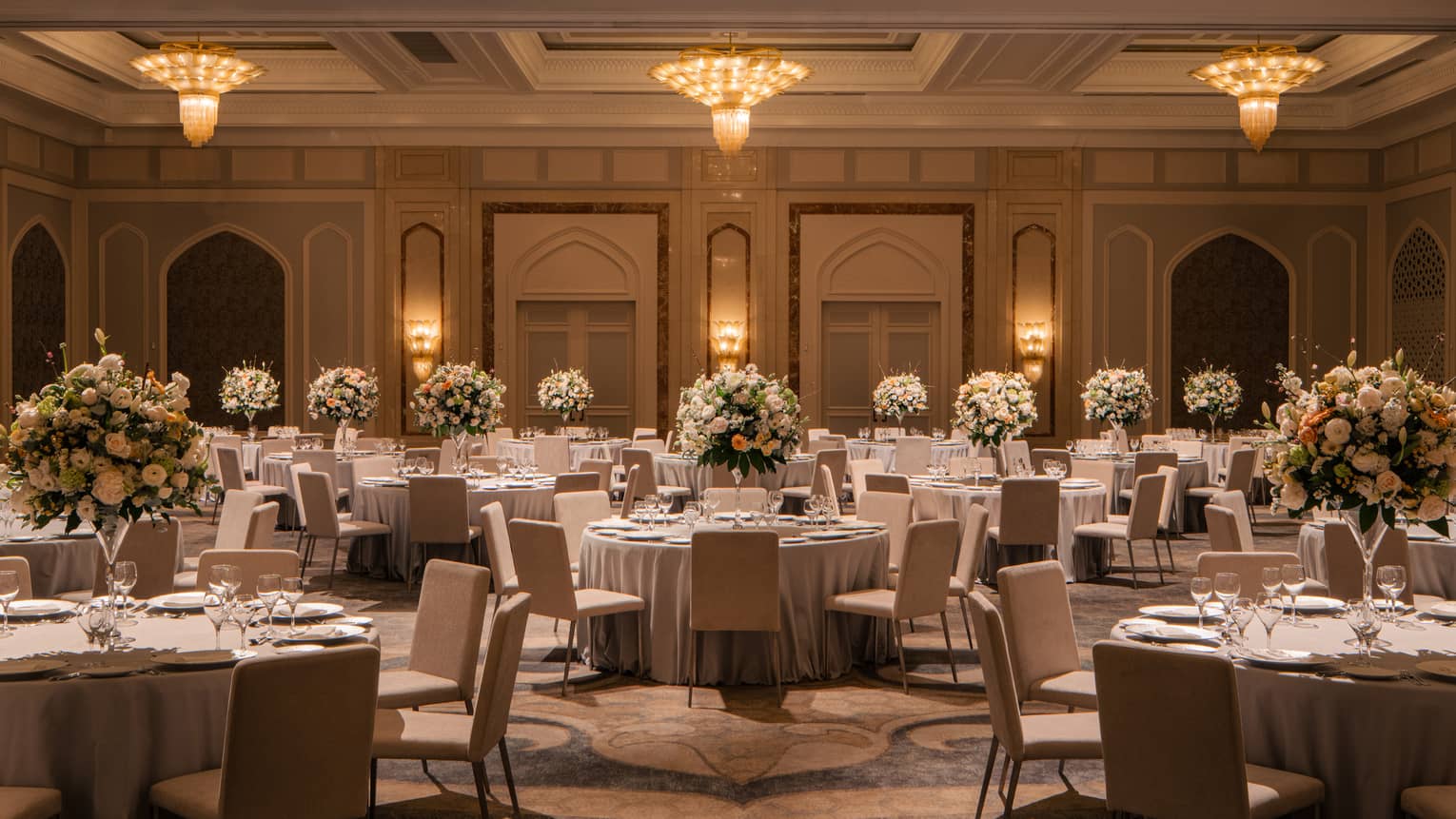 An elegant ballroom with chandeliers, ornate ceilings and banquet tables topped with large floral bouquets