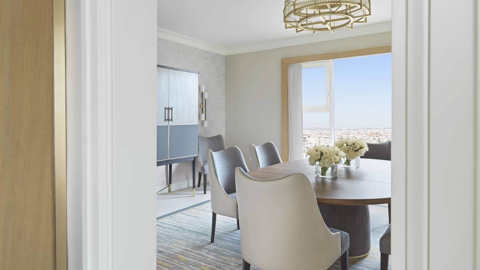 A doorway shot of a brown circular table surrounded by plush white chairs under a golden modern chandelier. 
