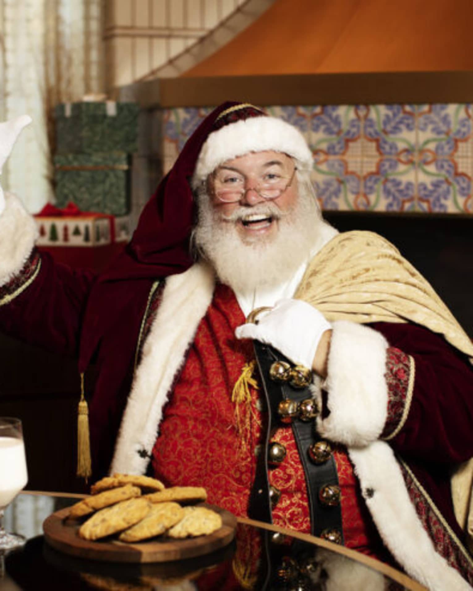 Santa Claus sitting at a table with a plate of cookies and glass of milk, and a fireplace in the background.