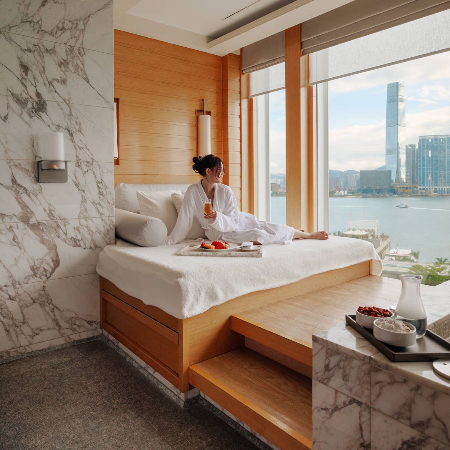 Woman in spa bathrobe sits on windowside chaise looking out to Victoria Harbour at Four Seasons Hotel Hong Kong