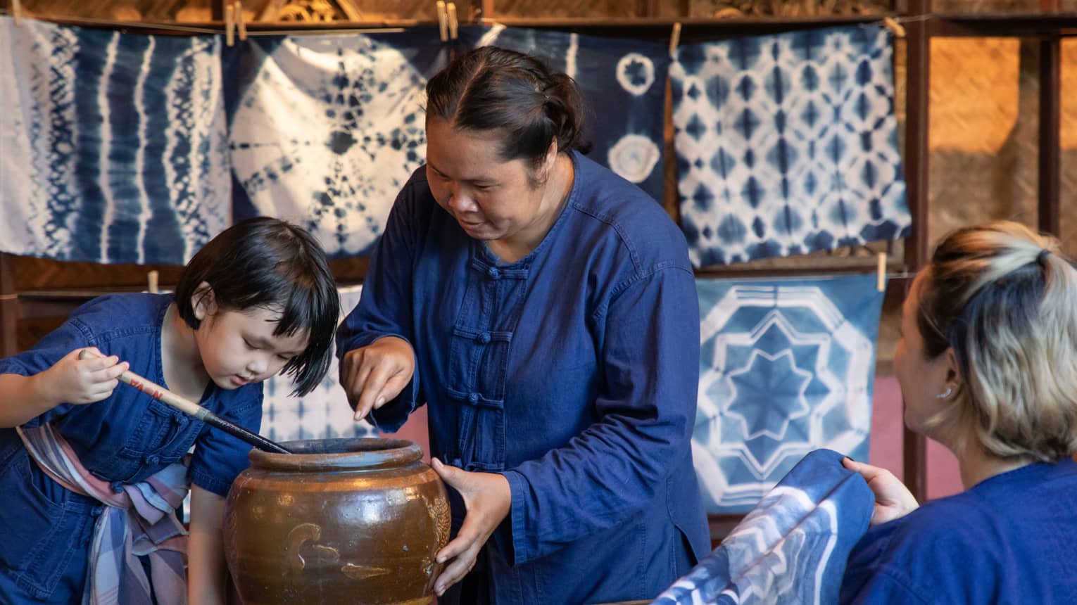 A woman teaching a child how to dye fabric using indigo, with tie-dyed cloths hanging in the background in a traditional workshop.