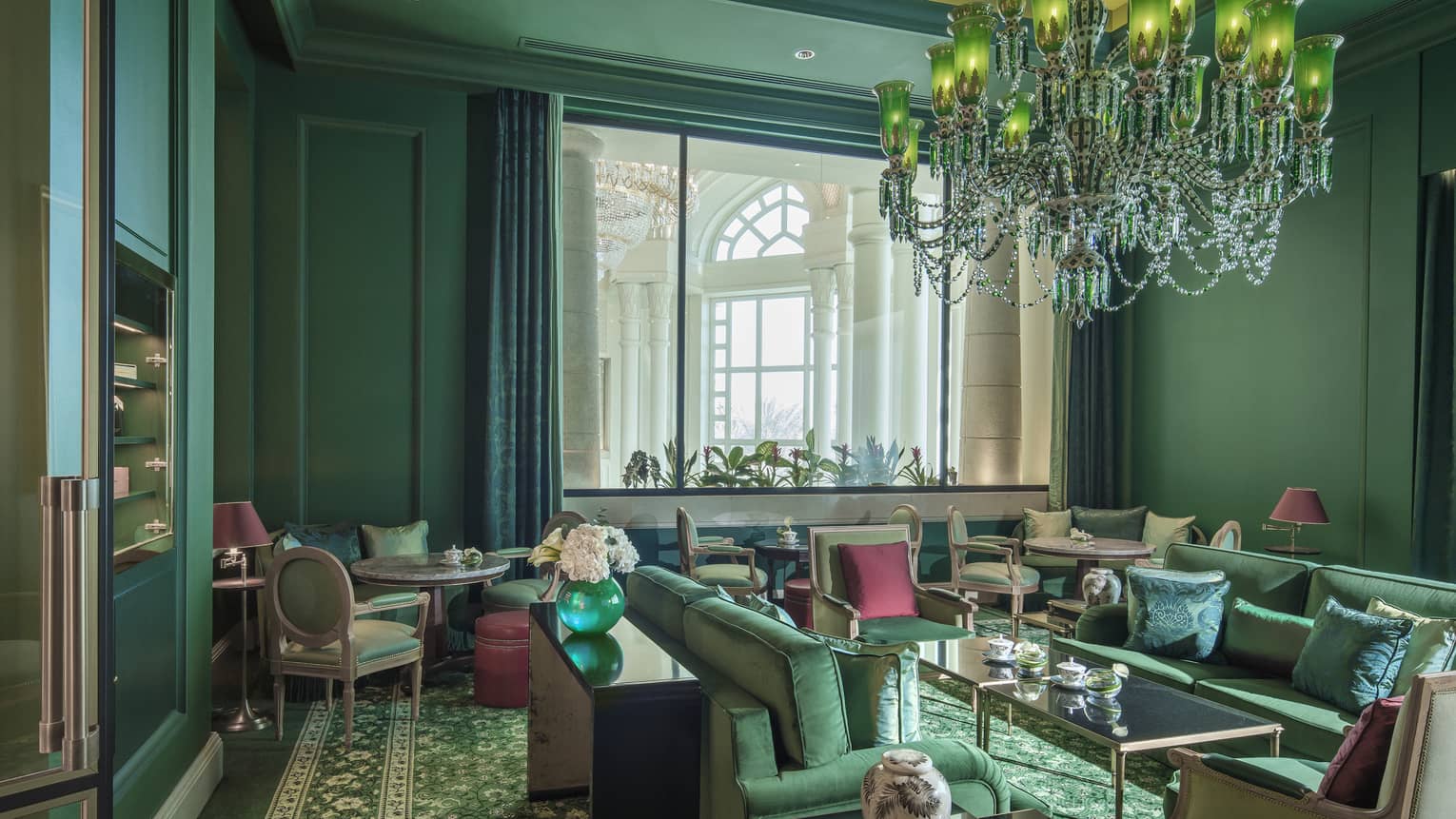 A forest green sitting room with over a dozen velveteen chairs, and a crystal chandelier. 