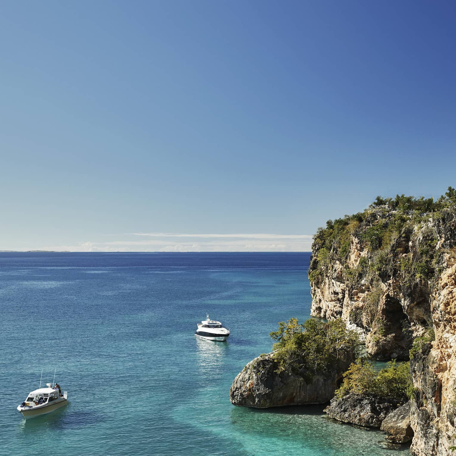 Long view of two cabin boats floating in the crystal-clear ocean beside towering rocky cliffs, a pale blue sky above.