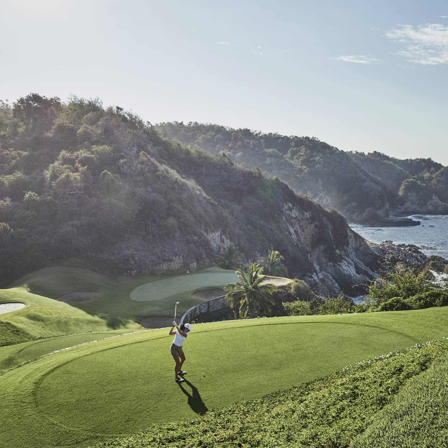 Golfer in mid-backswing, ready to strike toward a distant green at the base of an immense forested hill bordering the ocean.