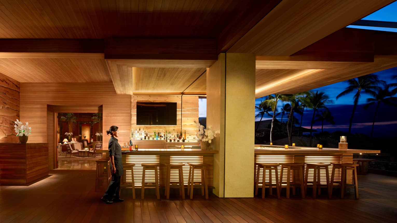 Server walks by wood NOBU Lanai restaurant bar with lights on indoor outdoor patio at night