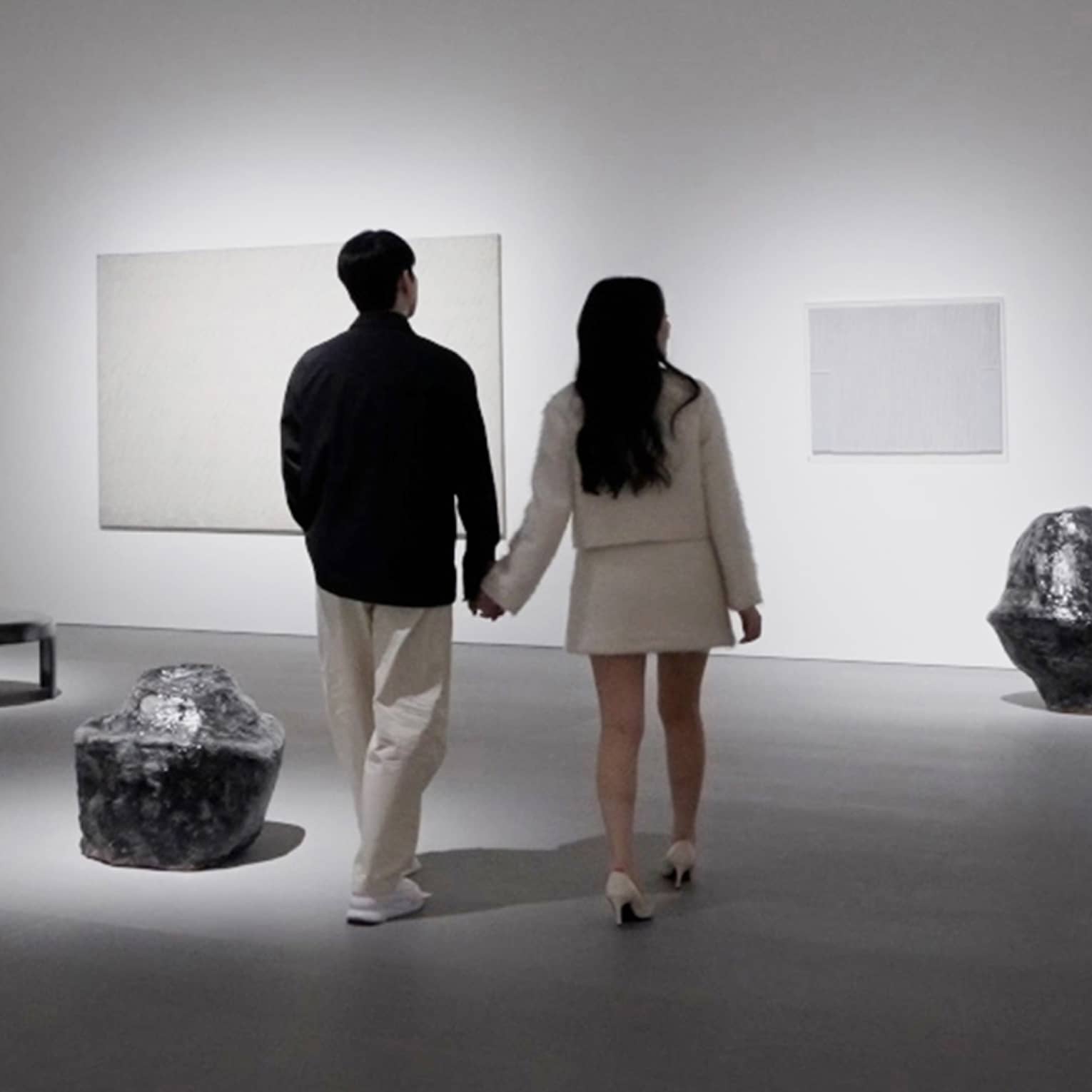 A couple holding hands walks through a modern art gallery with white walls, gray flooring and spotlights illuminating the art pieces