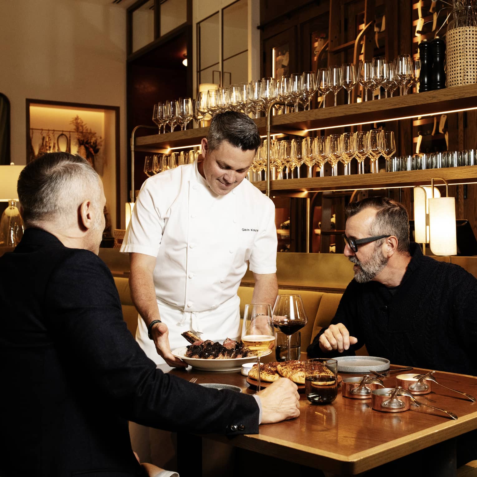 Chef brings a plate of food to two people seated at a table in front of a wood-and-brass shelf of wine glasses