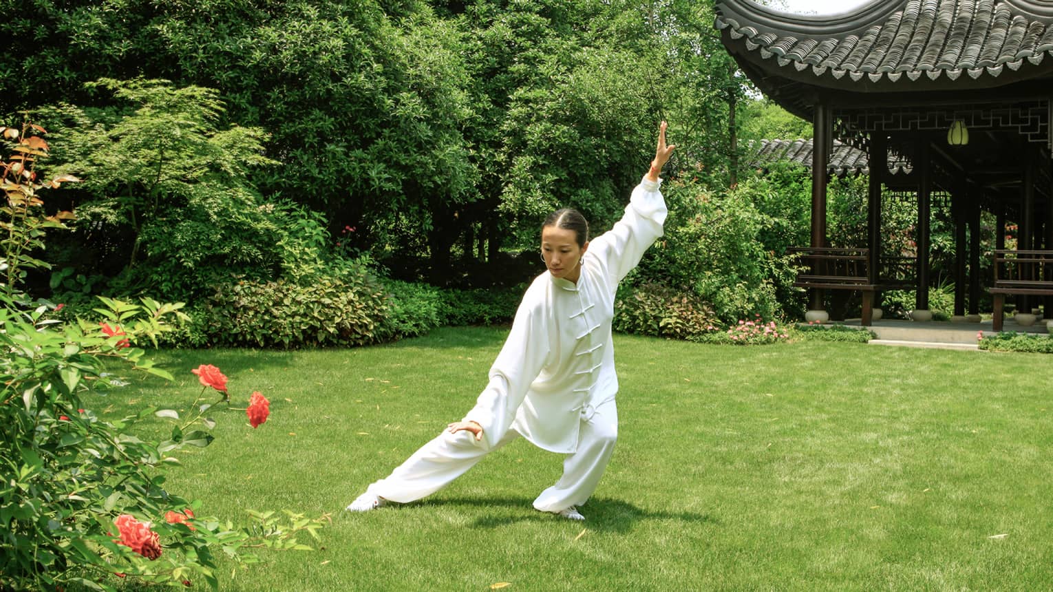 An instructor in a white tai chi outfit, arms and legs outstretched in a classic pose, in front of a pagoda-shaped gazebo.