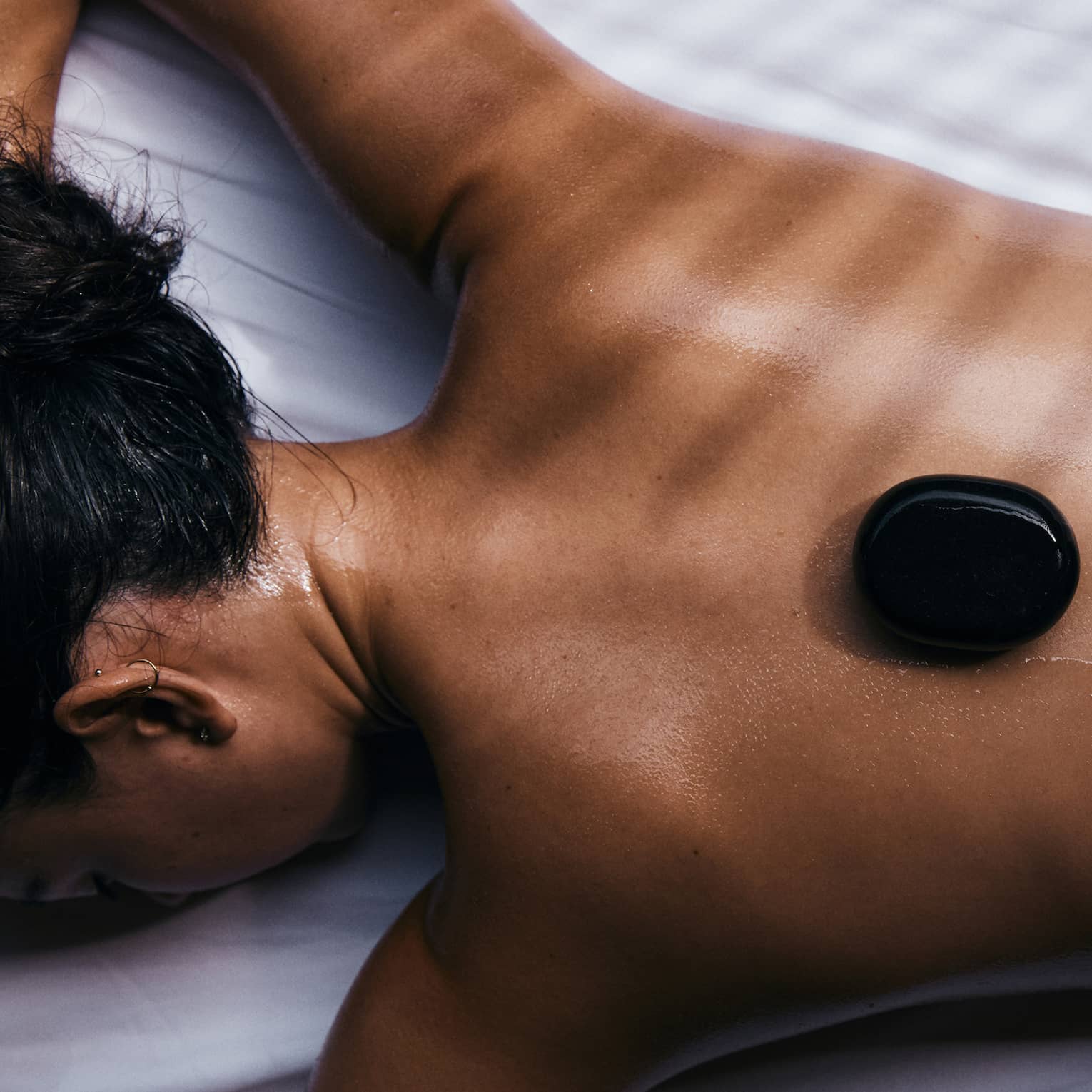 Close-up of a guest relaxing on their stomach on a bed of white linen with three black warming stones on their bare back.
