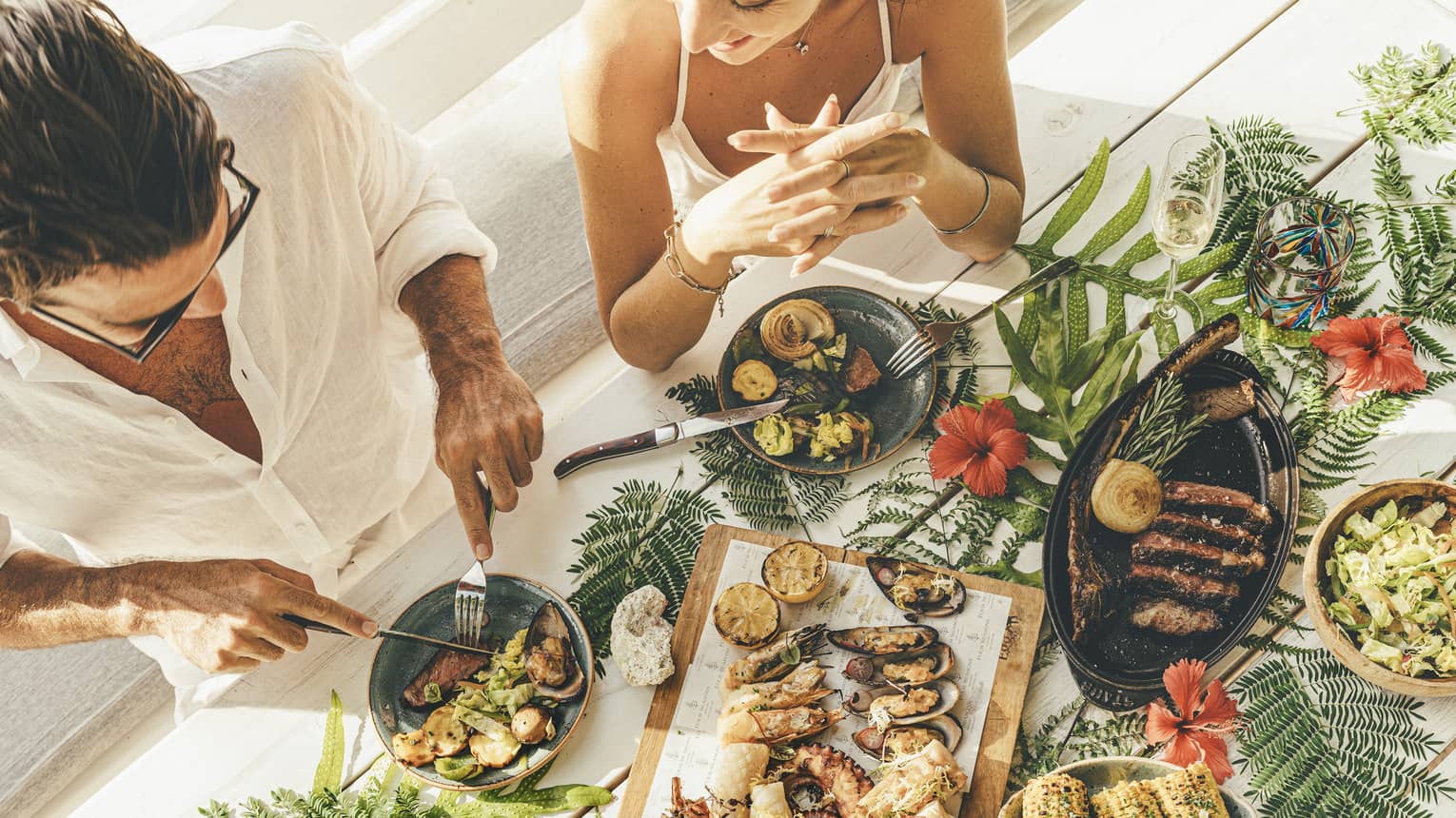 A couple at a table of seafood canapés, grilled meat, corn and champagne flutes, adorned with palm fronds and red hibiscus.