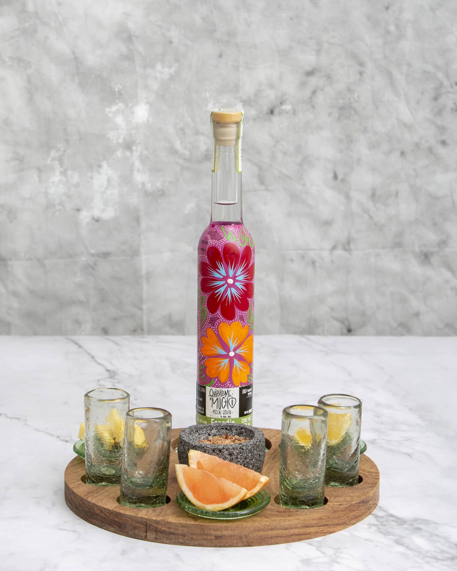 Brightly floral-painted mezcal bottle on wooden tray with four shotglasses, stone spice bowl and lemon and grapefruit wedges.