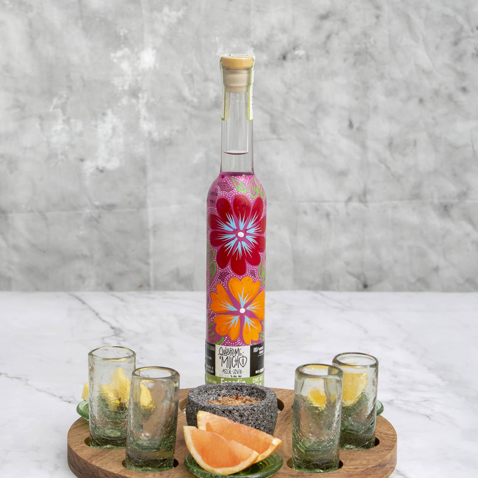 Brightly floral-painted mezcal bottle on wooden tray with four shotglasses, stone spice bowl and lemon and grapefruit wedges.