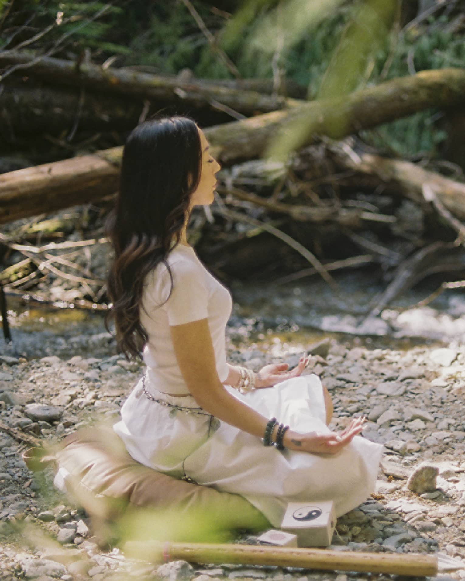 An instructor and a guest sit on yoga cushions as they peacefully meditate in a small clearing beside a brook in a forest.