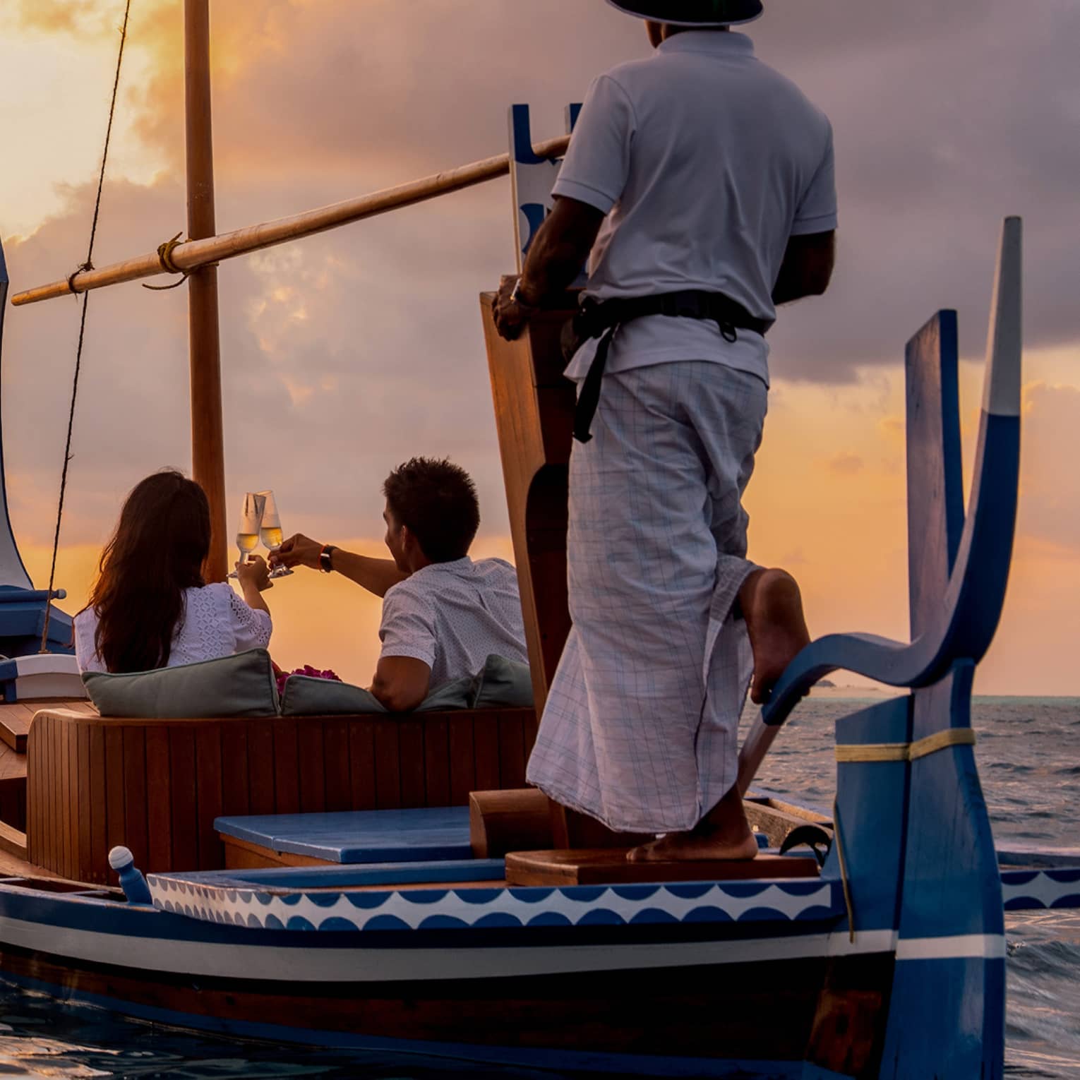 Rear view of two people seated in a dhoni boat, toasting with champagne, the boat handler steering into the golden sunset.