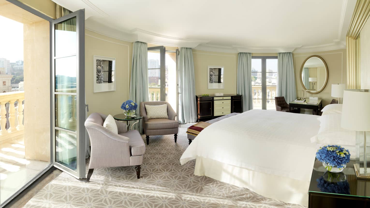Sun streams in from open balcony door to curved hotel room with bed, plush armchairs, mirrors