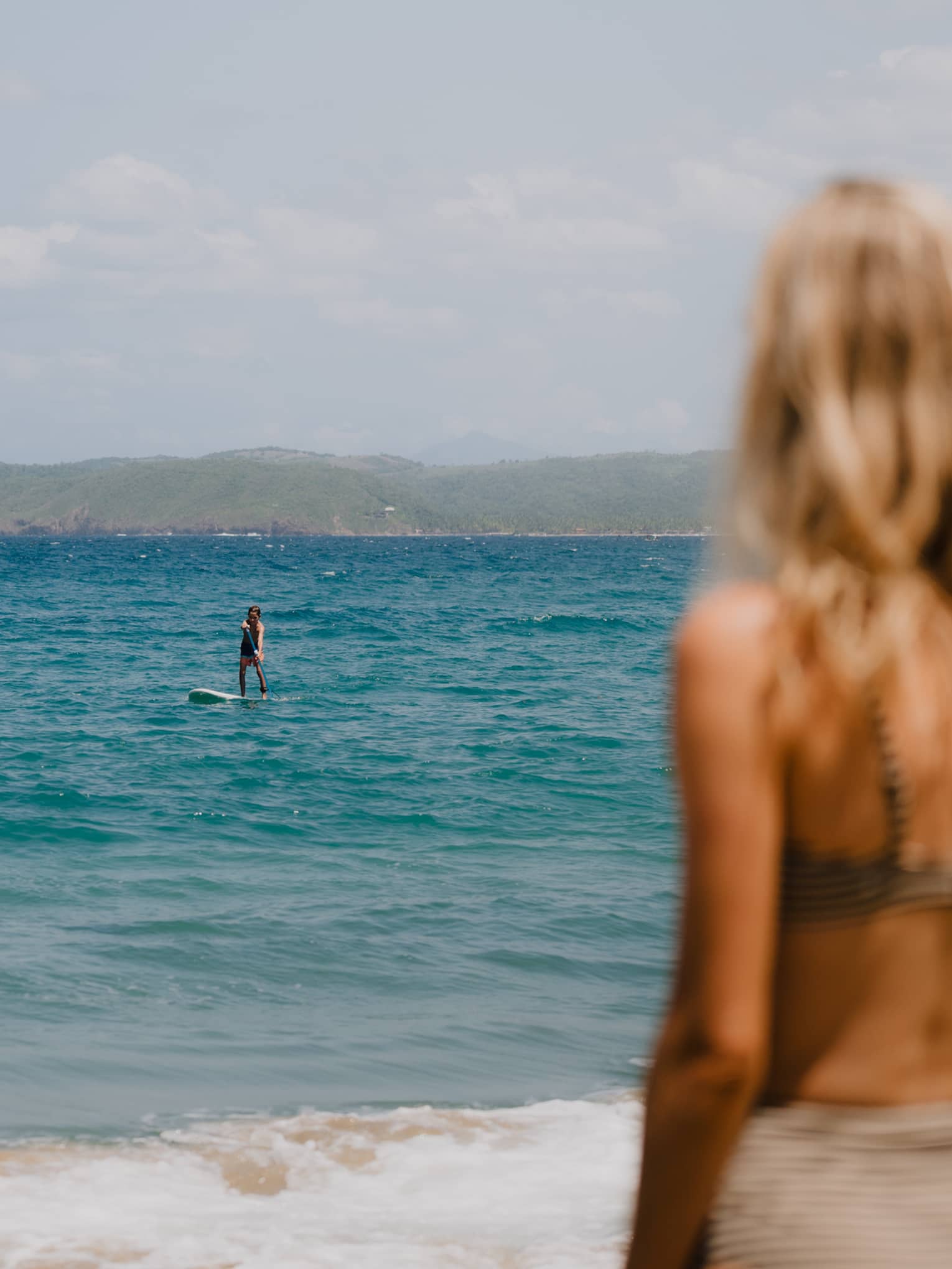 A person stands on a beach, facing the ocean, where other people are paddleboarding