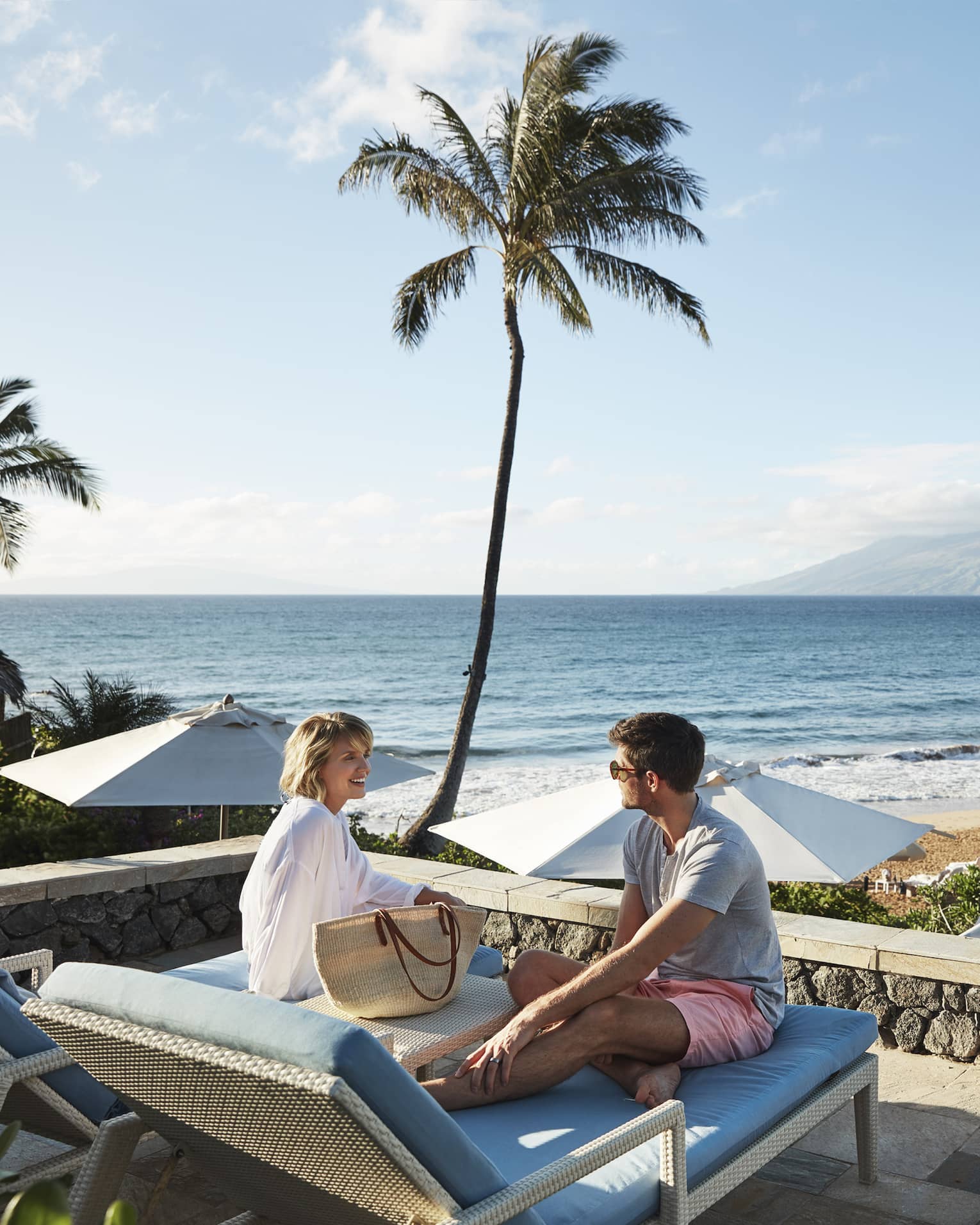 Man in shorts and smiling woman in beach shirt sit on edge of lounge chair, beach and ocean in background