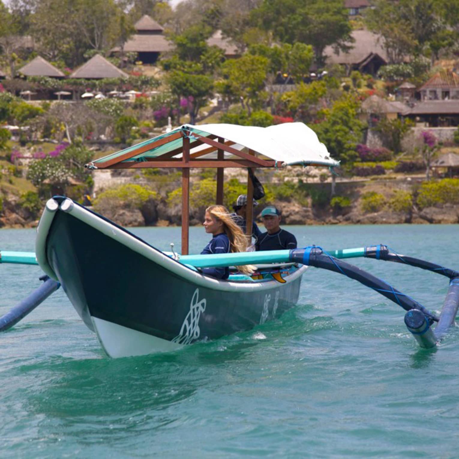 A guide sits at the stern of a covered outrigger canoe as two guests gaze at the water; behind, trees and huts dot the shore.