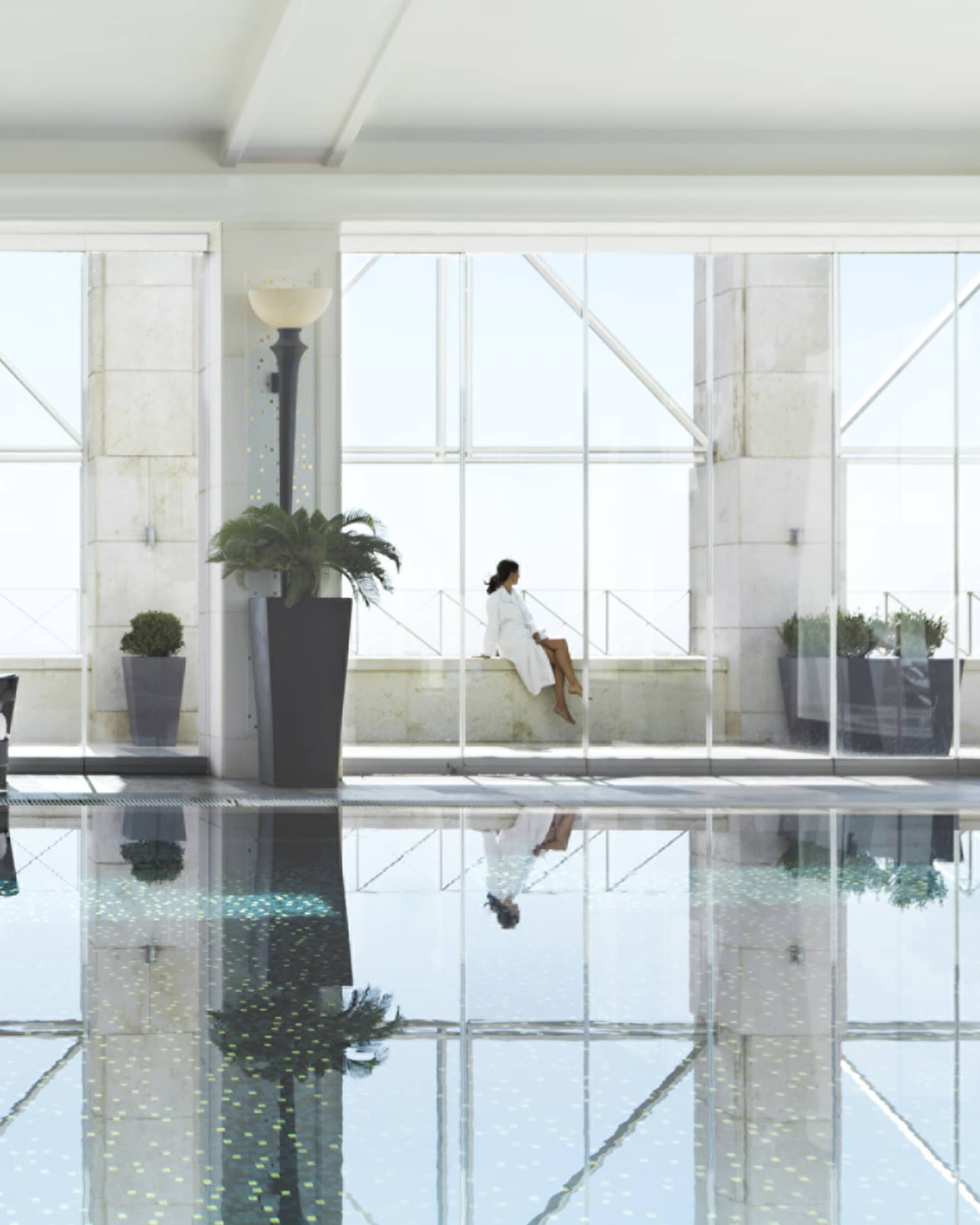 Indoor swimming pool reflecting three tall windows, woman in white bathrobe sits on ledge