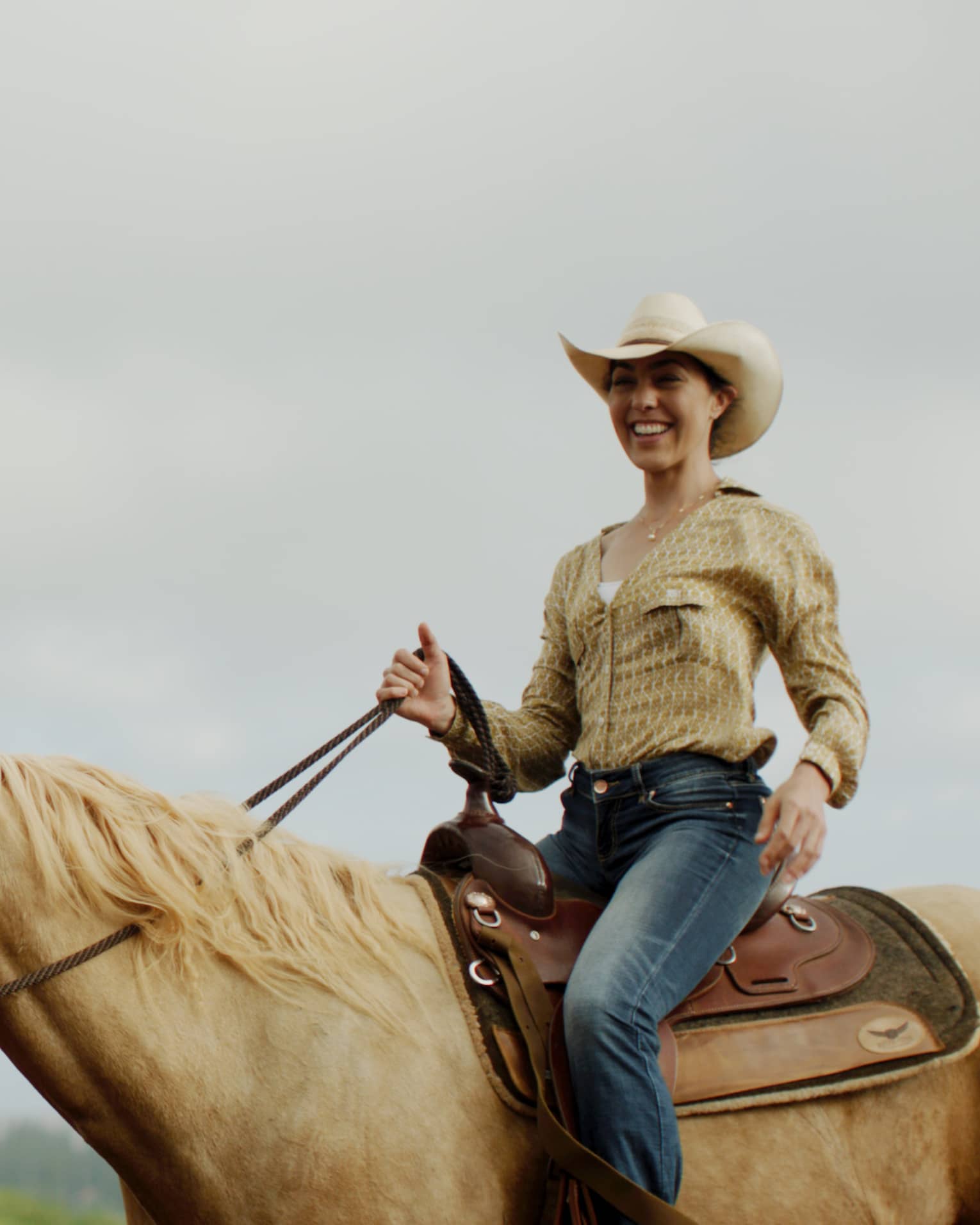 A smiling guest wearing a cowboy hat holds the reins while riding a tan horse under cloudy skies, forest in the distance.