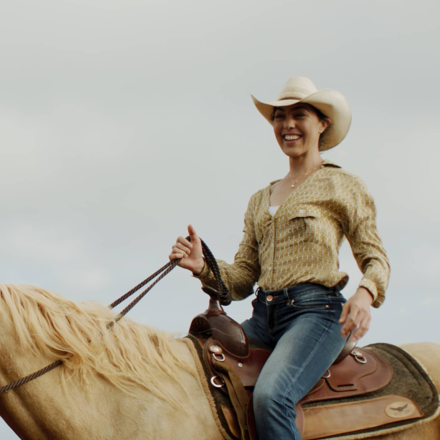 A smiling guest wearing a cowboy hat holds the reins while riding a tan horse under cloudy skies, forest in the distance.
