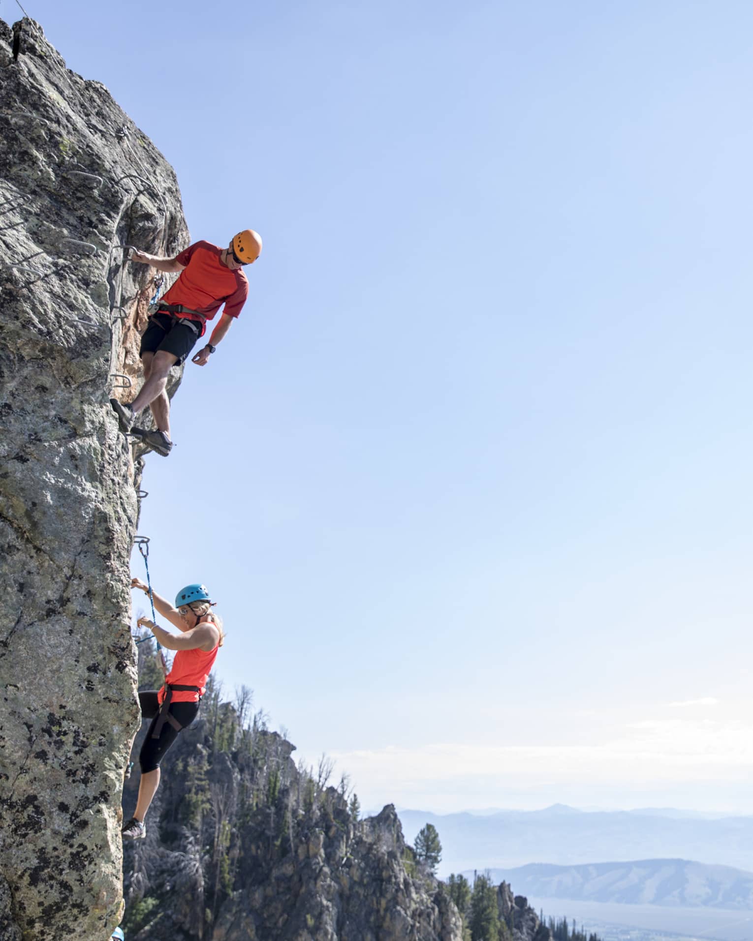 Man and woman climb steep cliff face with view of mountains in far distance 