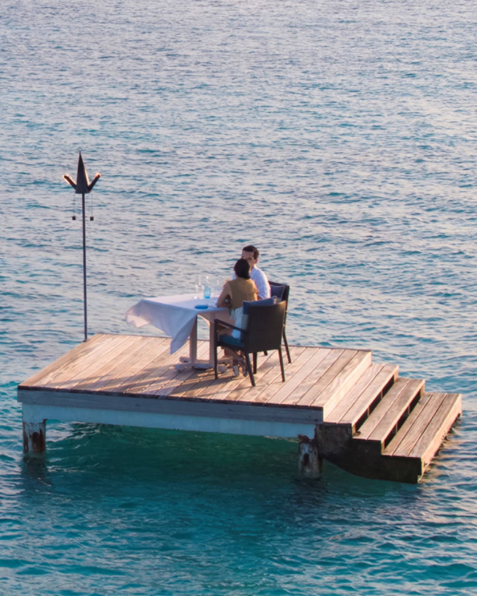 Man, woman dine at private table on small floating wood platform in middle of blue lagoon