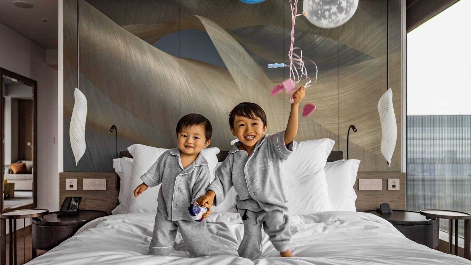 Two young children in grey pyjamas hold hands while standing on a bed in a bright room; one smiles and holds a clear balloon.