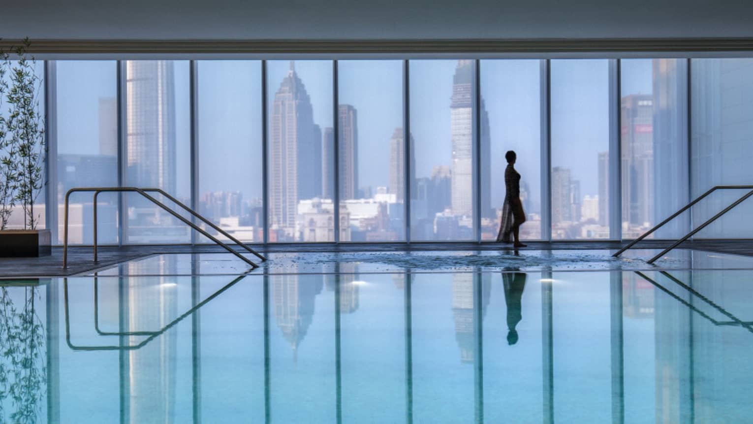Silhouette of woman walking by blue indoor swimming pool, floor-to-ceiling window