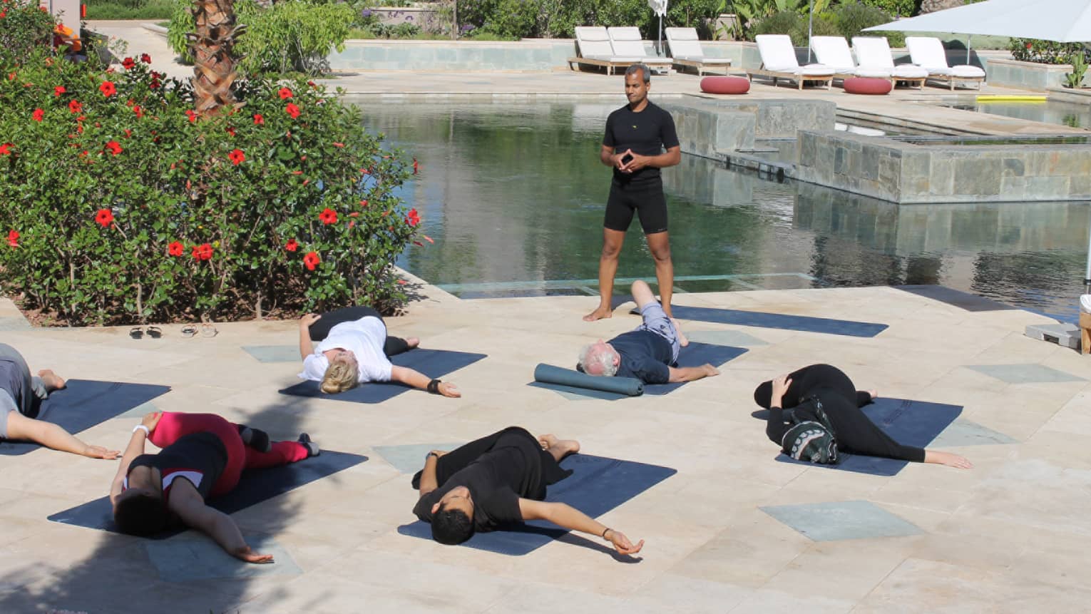Fitness class on patio, instructor stands over people stretching on yoga mats
