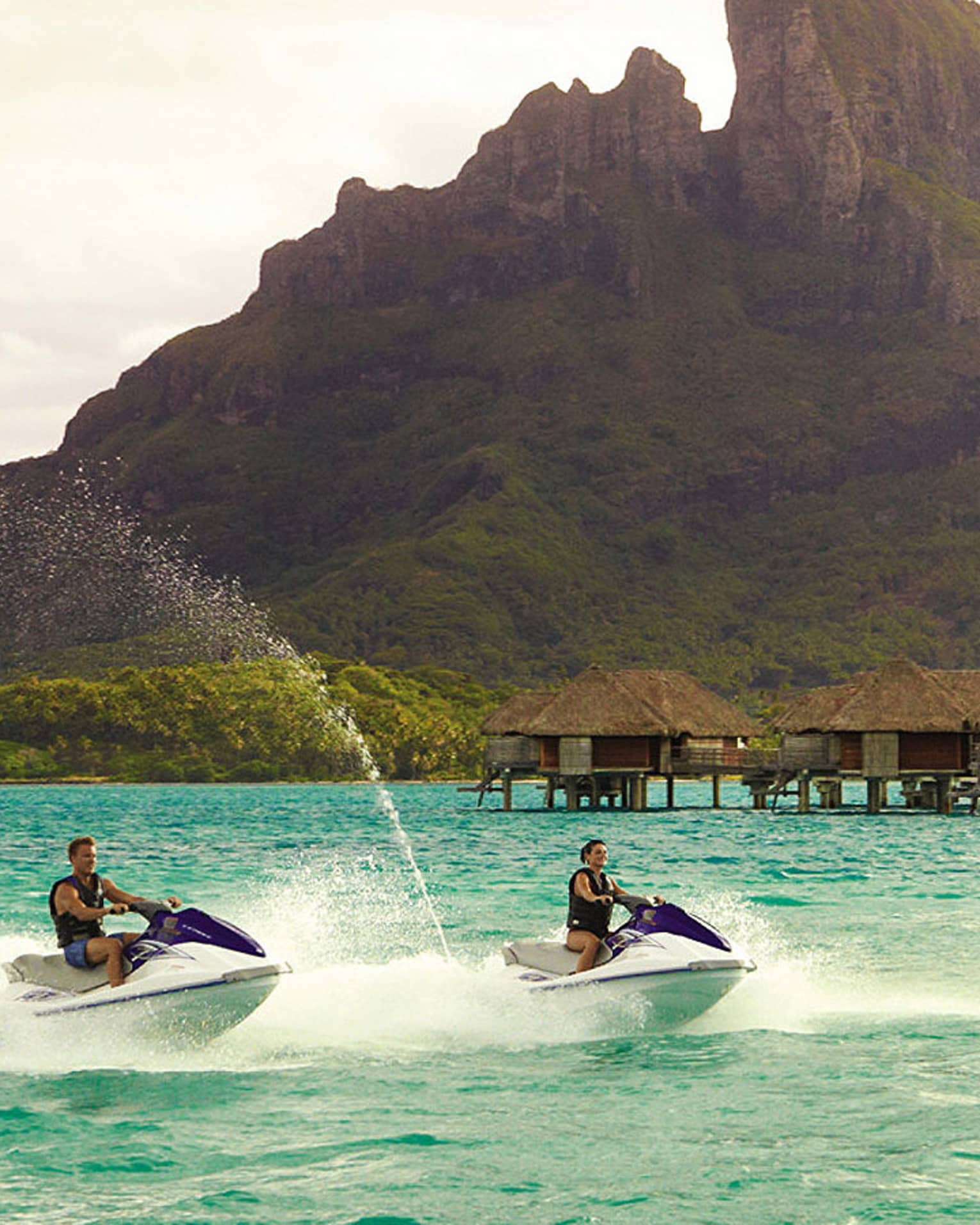 Two jet skis ride along lagoon past row of overwater bungalows, Bora Bora mountain