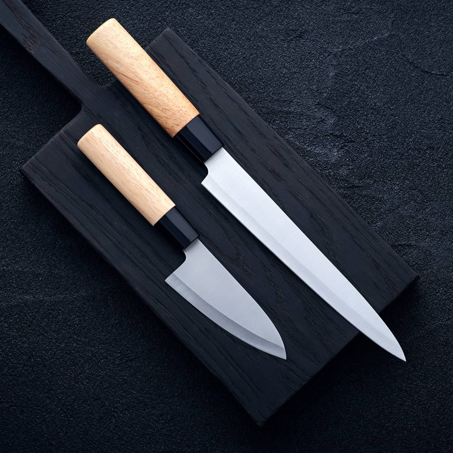 Two extremely sharp wood-handled knives, one long, one short, laid out on a black cutting board on a dark surface.