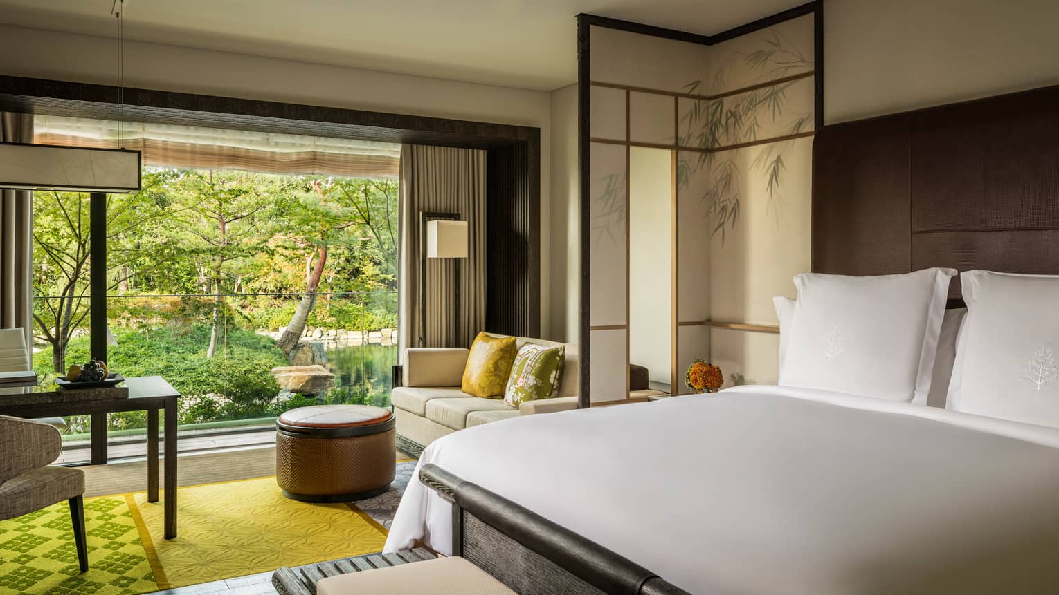 Guest room with a king bed, sofa with green cushions, round ottoman and large window overlooking a pond garden