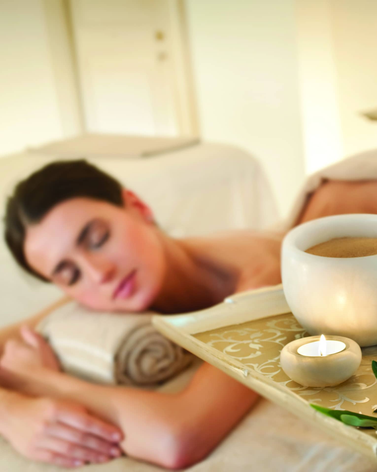 Spa staff holds tray with tealight candle, perfume, marble bowl near woman lying on massage table