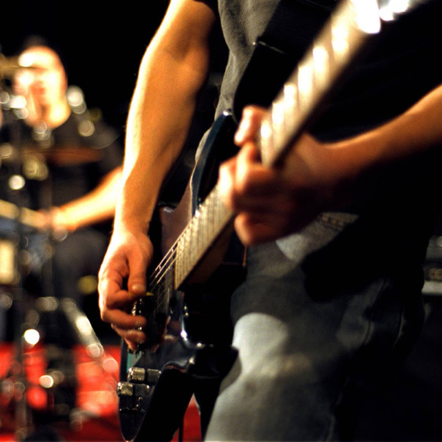 Close-up of musician's arms and hands playing an electric guitar with drumset in the background.