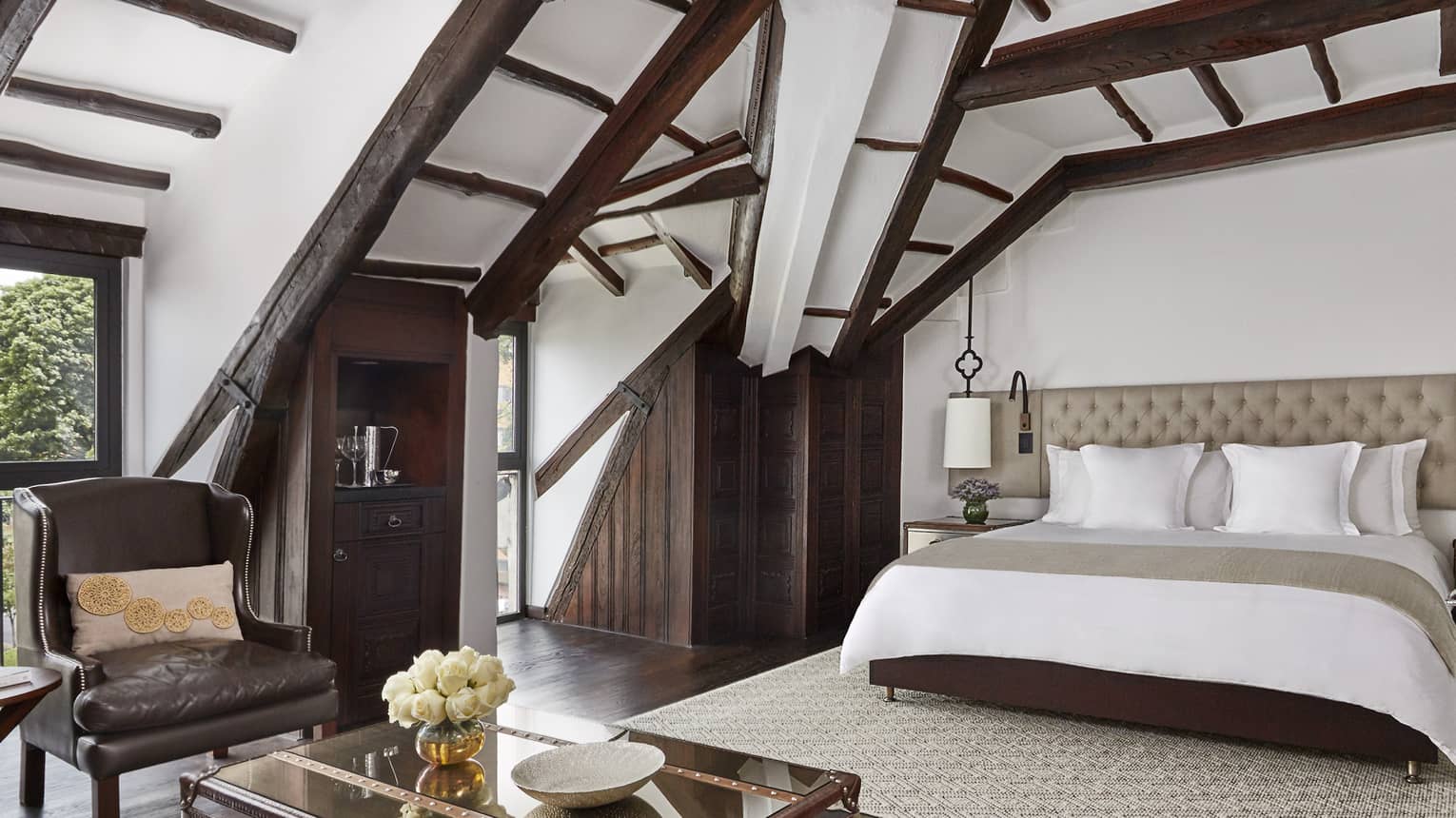 Sloped beam loft ceiling over hotel bed with padded headboard, brown leather armchair, table with flowers