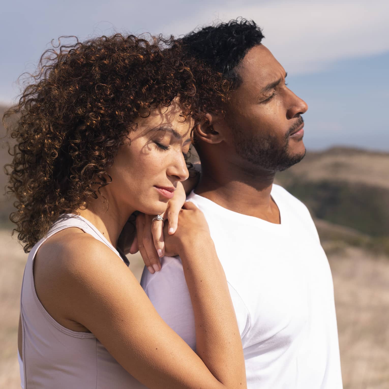 Two people both wearing white t-shirts stand together in a field with their eyes closed, one with their arms on the other's shoulders