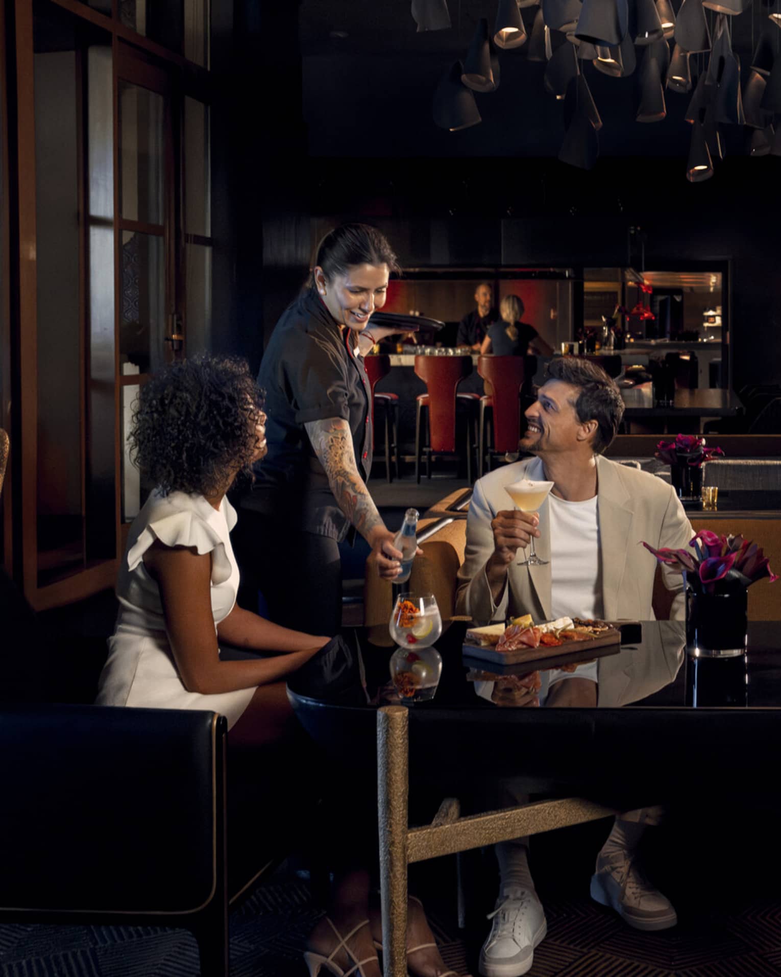 Two people wearing light-coloured cocktail attire sit on benches around a black table as a waitress brings them beverages