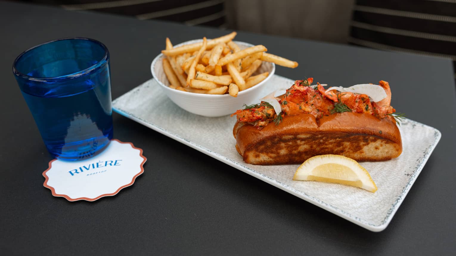 A rectangular plate holds a sandwich, a side of fries and a lemon slice. A blue tumbler sits next to the plate.