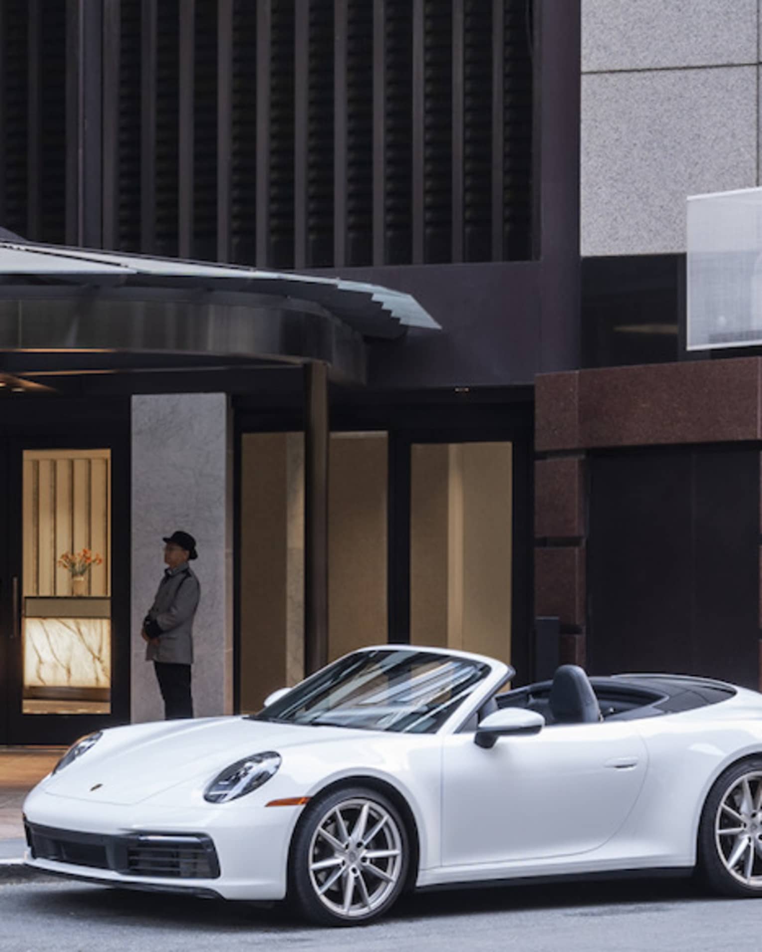 A white luxury sports caris parked in front of the massive Four Seasons hotel entrance, which is clad in pale grey granite.