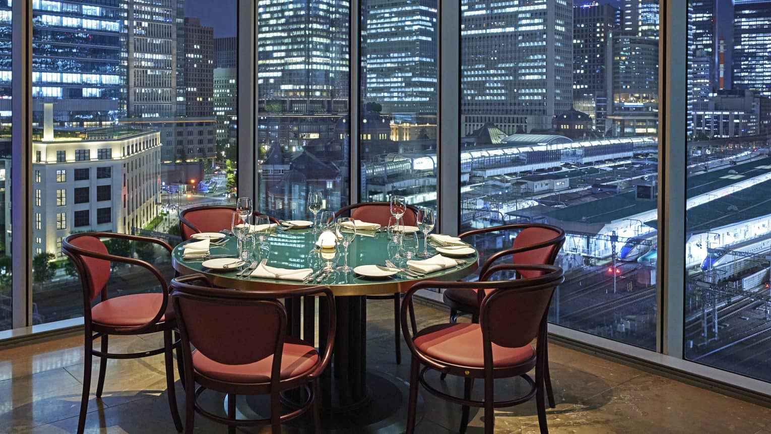 Luxury boutique hotel dining setup with a round table elegantly set for guests, offering a panoramic city view at night through tall glass windows