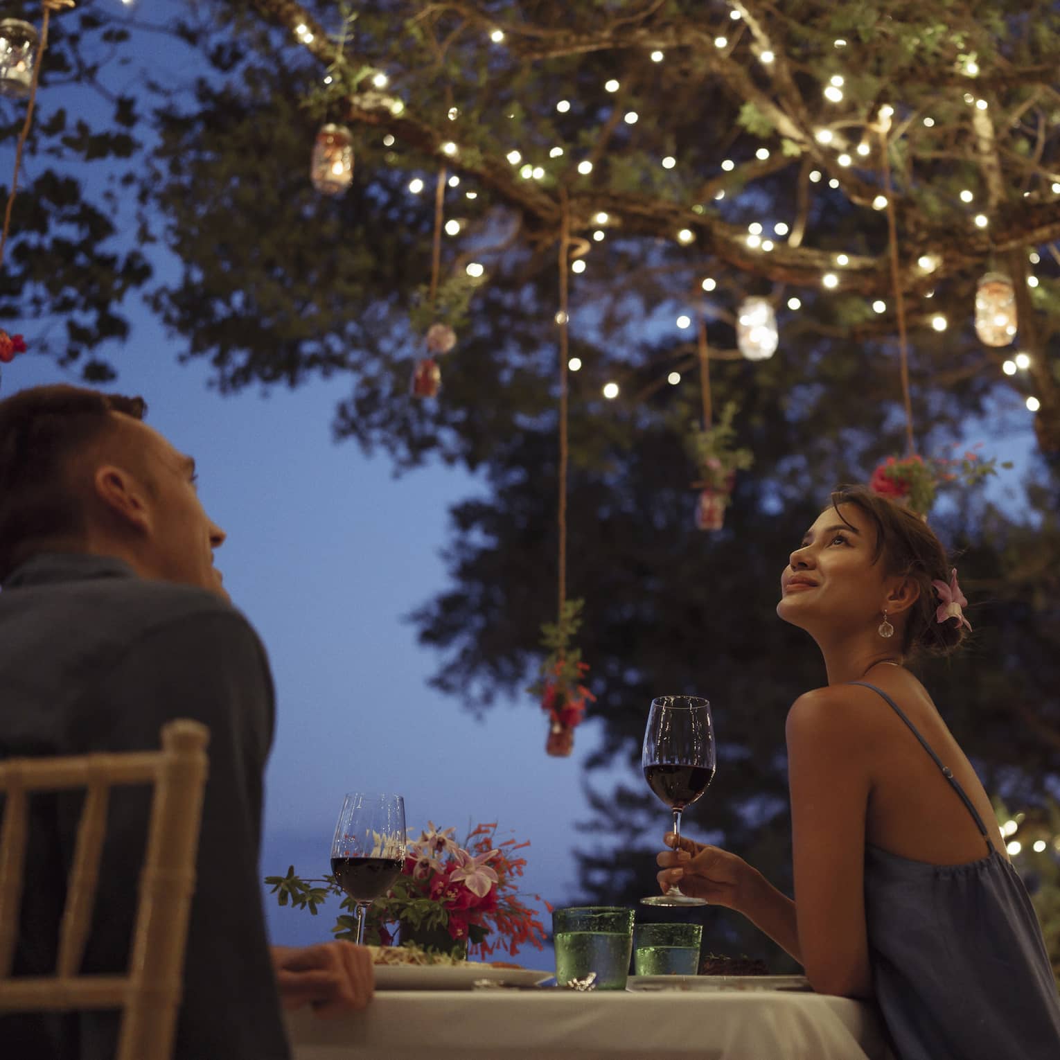 A smiling couple having a romantic dinner under trees covered in string lights and jars of flowers hanging from the branches.